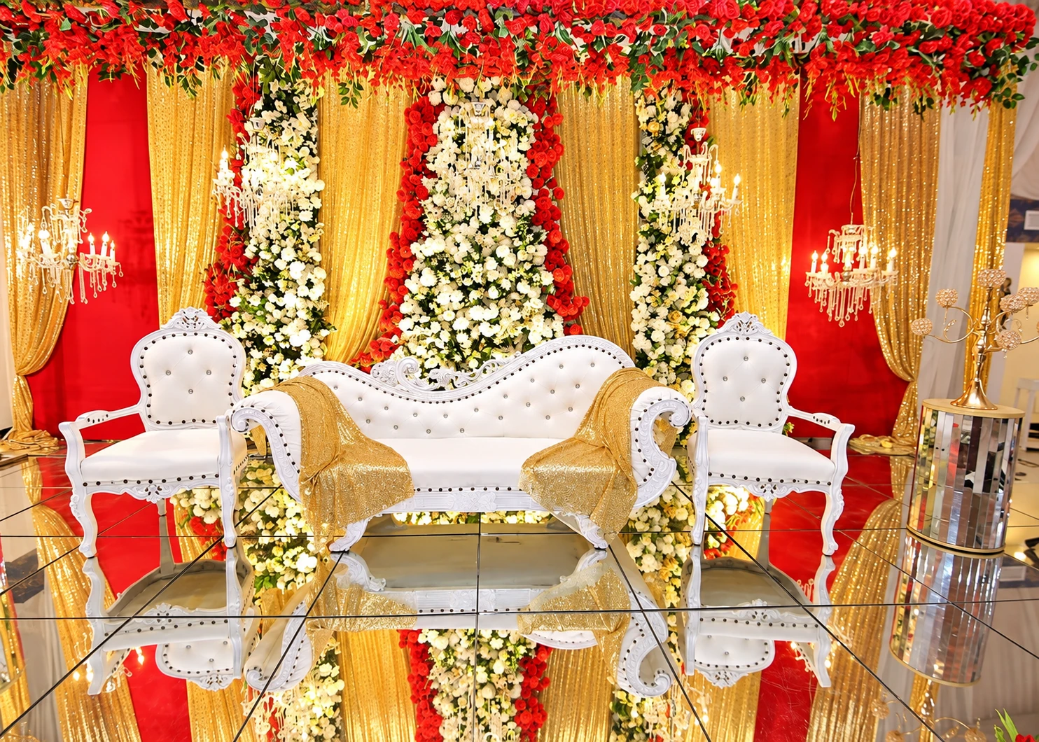 Red and gold Baraat stage with white tufted sofa on a mirror floor